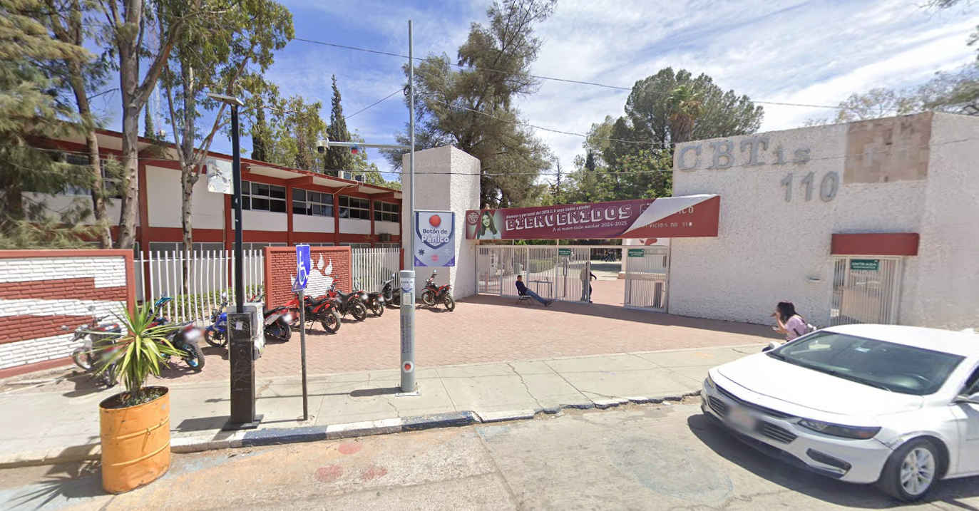 Fotografía en la que se muestra una explanada. Al fondo hay una reja de color blanco y sobre ella hay una placa roja que dice BIENVENIDOS. Del lado derecho de la reja hay un muro blanco que en letras grandes grises dice Cebetis 110.
